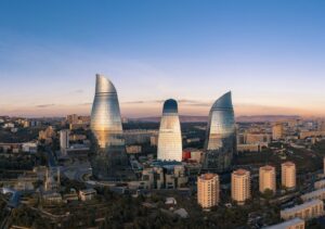 A panoramic sunset view of the Baku skyline in Azerbaijan, featuring the three iconic Flame Towers illuminated against a soft orange and blue sky, with the Caspian Sea and city buildings in the foreground.