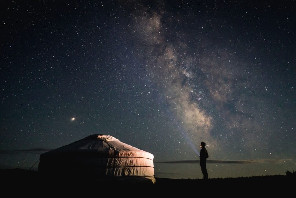 A silhouette of a person standing under a vast, brilliant Milky Way galaxy at night next to a traditional Mongolian yurt (ger).