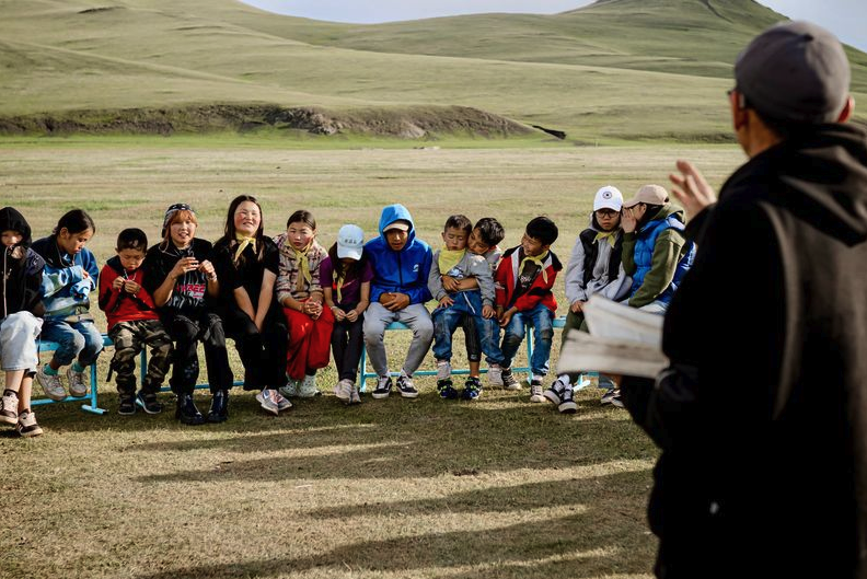 A group of children and adults sitting together outdoors in a grassy Mongolian landscape, participating in a community gathering or educational session.