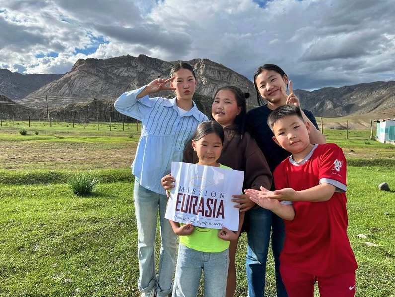 Four smiling children standing in a grassy field in the Eurasian Steppe, with one child holding a Mission Eurasia" sign and mountains in the background.