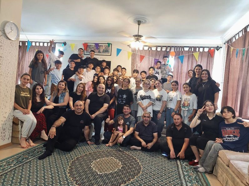 A large, diverse group of people of various ages, including children, teenagers, and adults, posing together for a group photo in a brightly lit indoor living room. Many are smiling, and the room is decorated with colorful festive banners hanging from the ceiling.