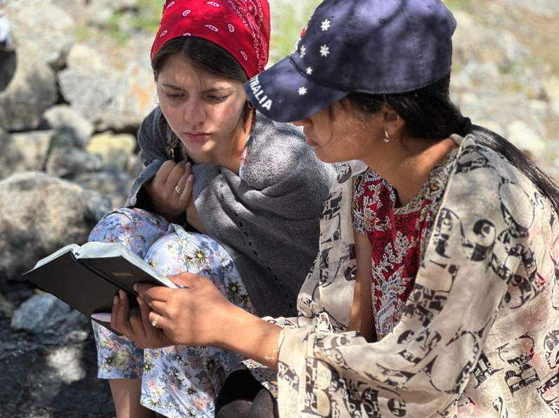 Two women in Tajikistan sitting outdoors and focused on reading a book together.