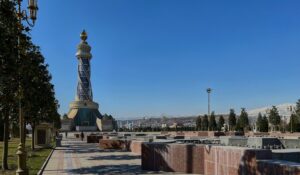 A wide view of the Ismoili Somoni Monument and a tall ornamental tower in a public square in Dushanbe, Tajikistan, under a clear blue sky.