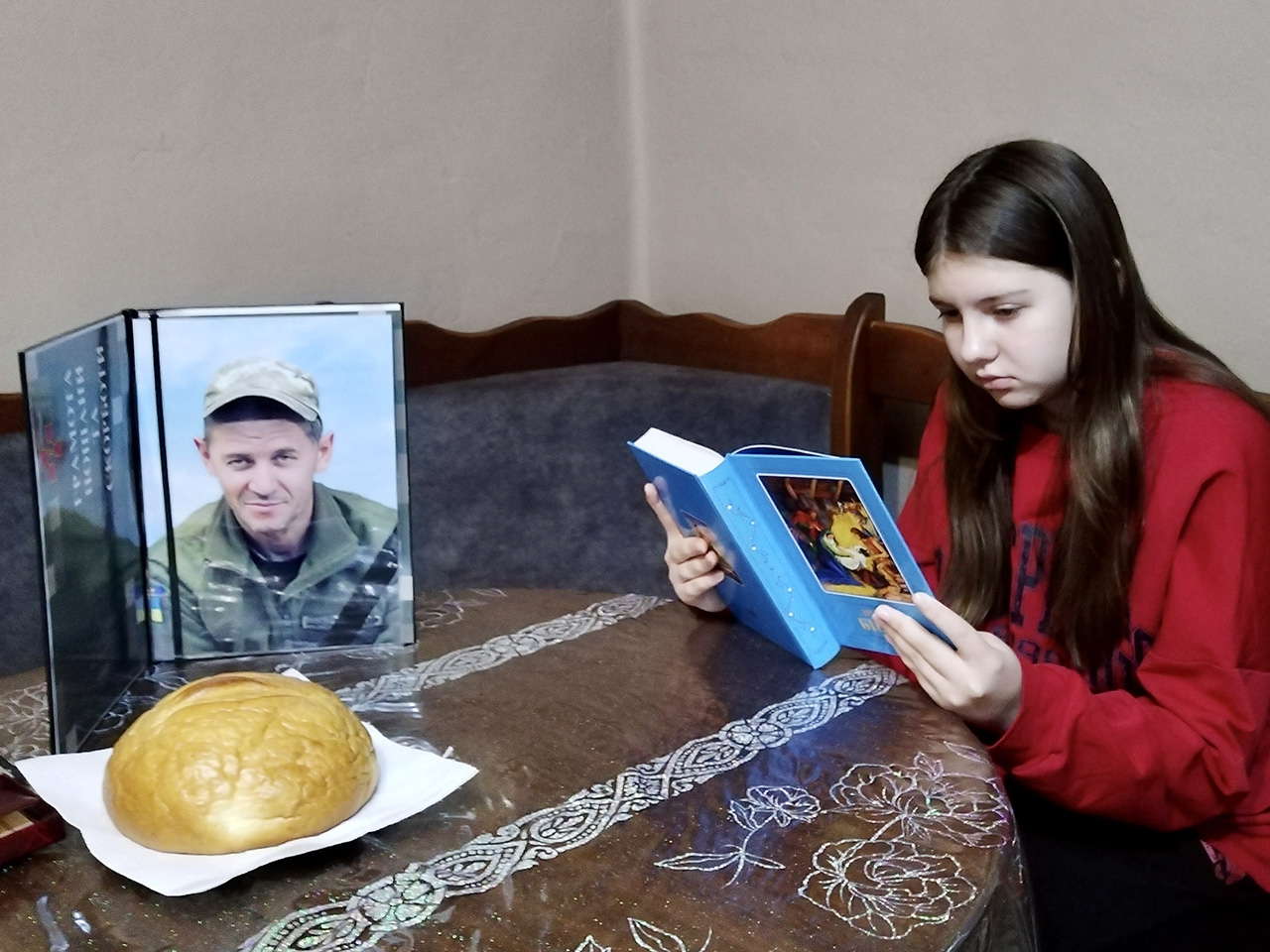 A young girl in a red sweatshirt sits at a wooden table, looking down sadly at an open book. On the table sits a framed photograph of a man in military uniform and a single loaf of bread.