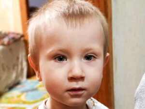 A close-up portrait of a young boy with light brown hair looking directly into the camera with a serious, slightly sad expression, highlighting the emotional toll on orphaned children.