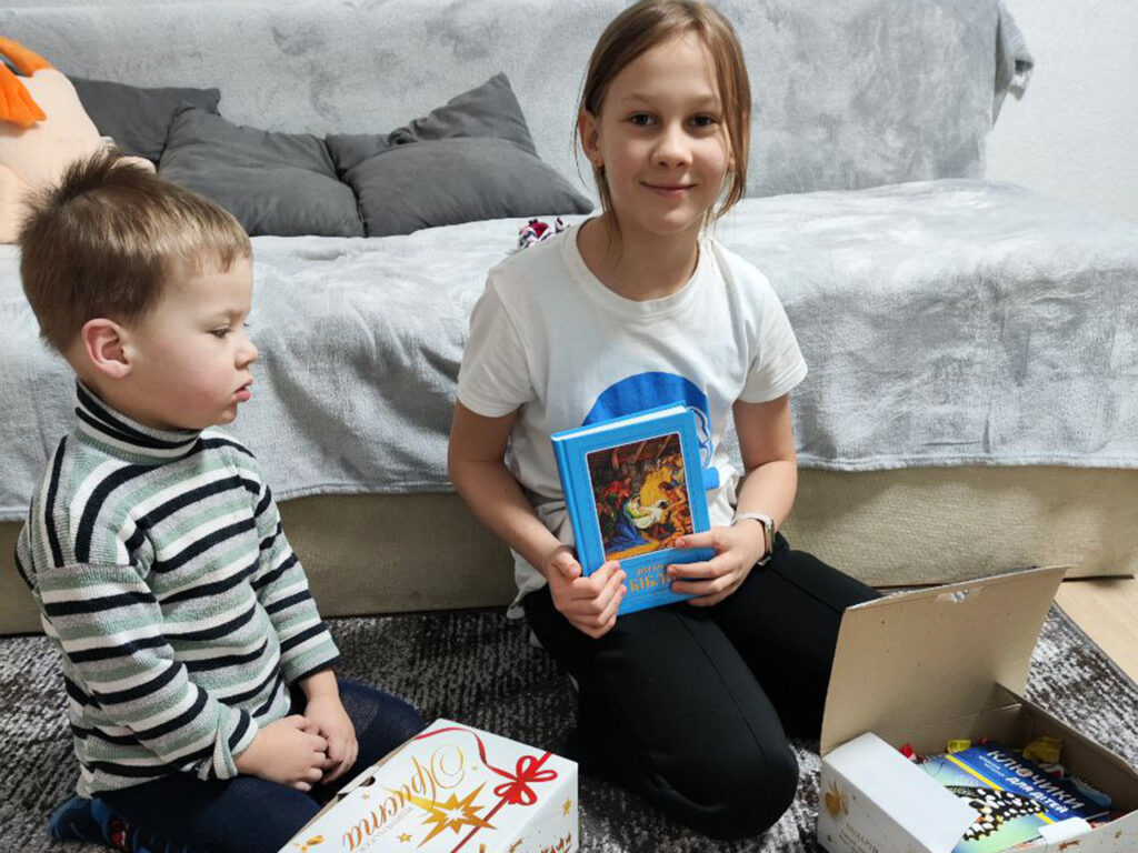 A young girl smiles while holding a blue book, sitting next to a young boy as they open a "Gift of Hope" care package filled with books and supplies.