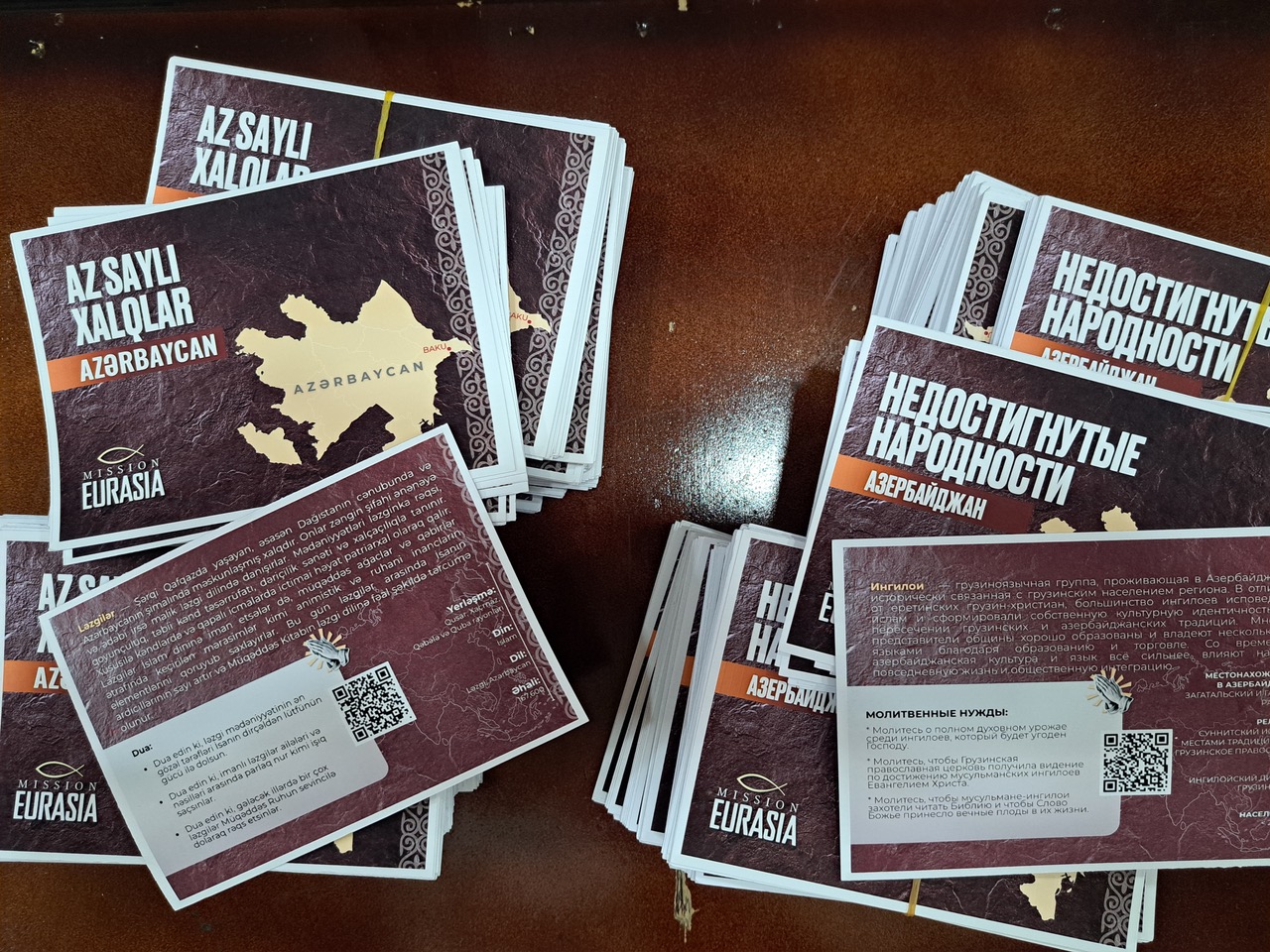 Stacks of Mission Eurasia ministry booklets and prayer cards written in Azerbaijani and Russian, featuring the organization's logo and QR codes.