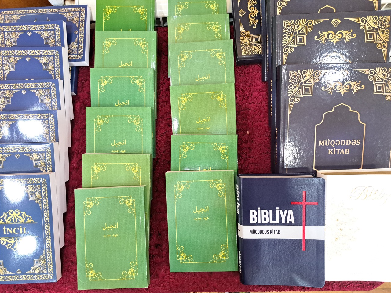 A collection of Bibles and Scripture booklets in various languages, including "Bibliya" and "İncil," displayed in stacks on a red surface.