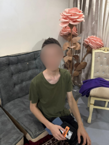 A young man in a green t-shirt sitting on a grey couch in front of large, decorative pink paper flowers.
