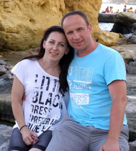 A smiling man in a light blue shirt stands alongside his wife in front of a rocky shoreline background.