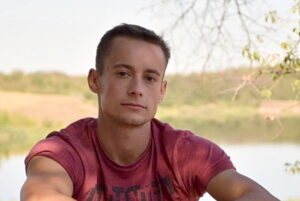 A close-up portrait of a young man, Viktor Maniushkin, wearing a maroon shirt and looking directly at the camera, with a soft-focus background of trees and a lake on a sunny day.