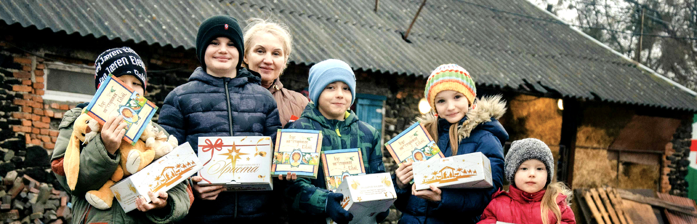 A group of smiling children and a woman standing outside a rustic building, happily holding up decorated Christmas gift boxes and books from Mission Eurasia.