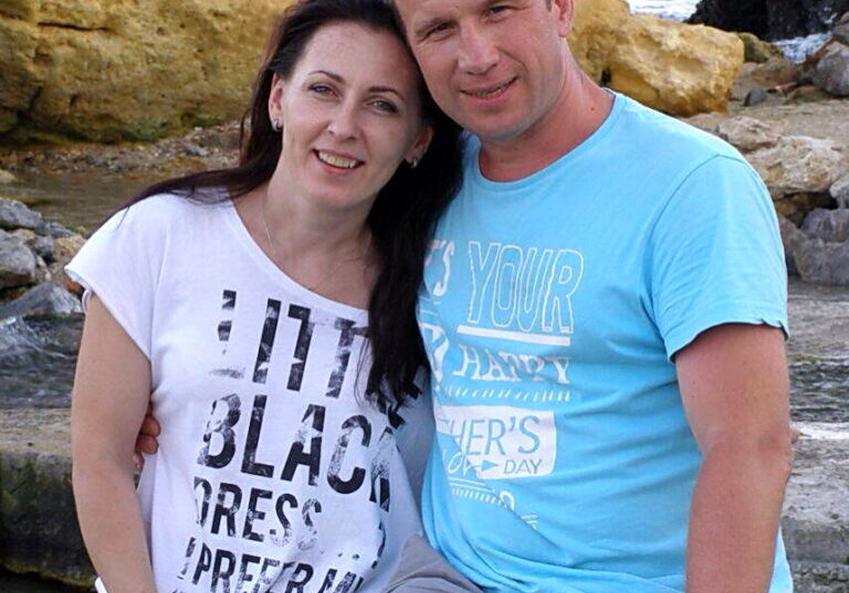 A smiling man in a light blue shirt stands alongside his wife in front of a rocky shoreline background.