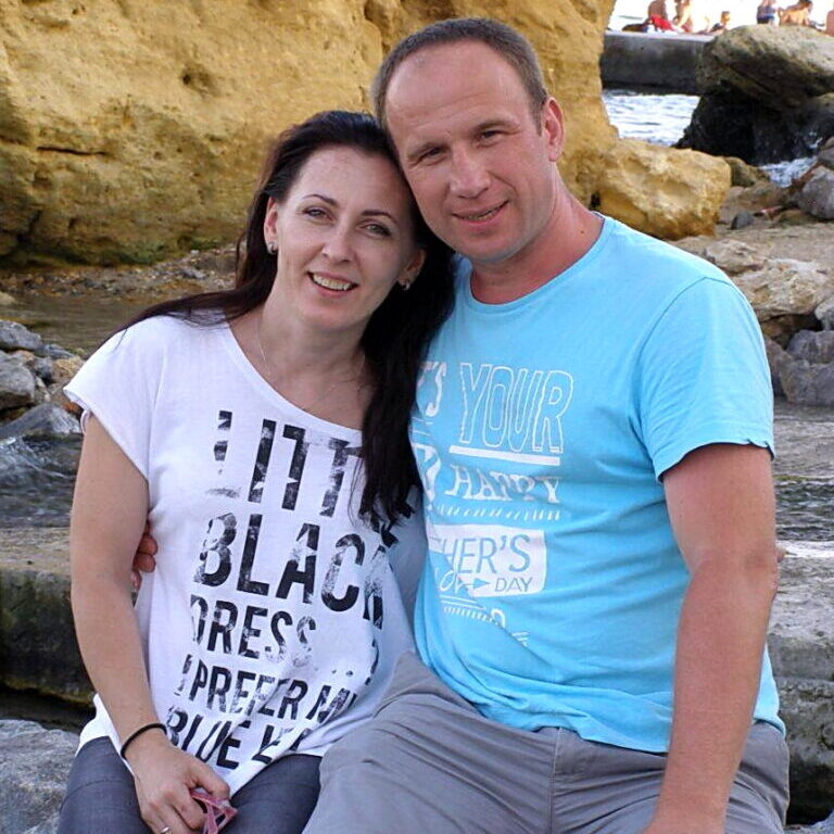 A smiling man in a light blue shirt stands alongside his wife in front of a rocky shoreline background.