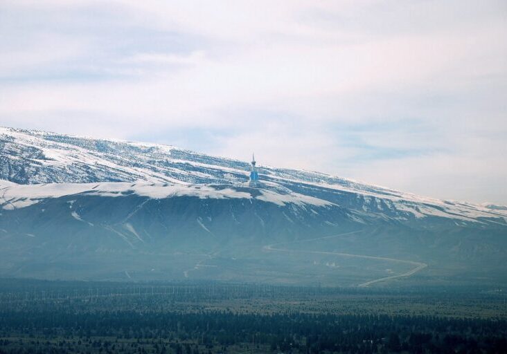 A wide landscape view of a snowy mountain range in Turkmenistan under a hazy blue sky, with a sparse evergreen forest in the foreground.
