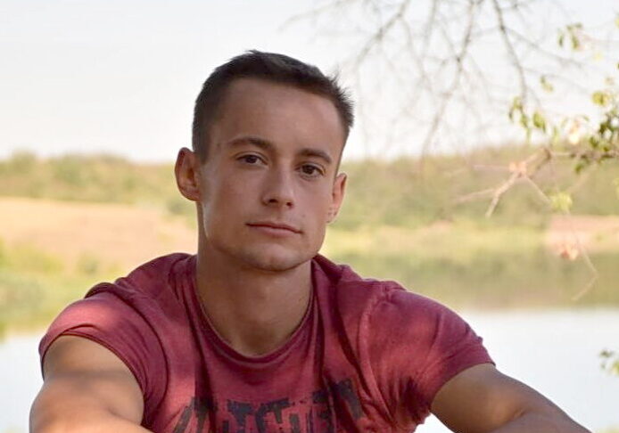 A close-up portrait of a young man, Viktor Maniushkin, wearing a maroon shirt and looking directly at the camera, with a soft-focus background of trees and a lake on a sunny day.