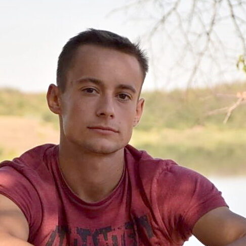 A close-up portrait of a young man, Viktor Maniushkin, wearing a maroon shirt and looking directly at the camera, with a soft-focus background of trees and a lake on a sunny day.