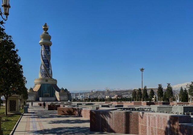 A wide view of the Ismoili Somoni Monument and a tall ornamental tower in a public square in Dushanbe, Tajikistan, under a clear blue sky.