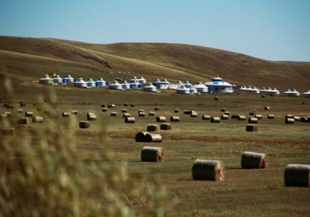 A wide view of the Eurasian Steppe featuring rolled hay bales in a field with a cluster of traditional white yurts and rolling hills in the background.